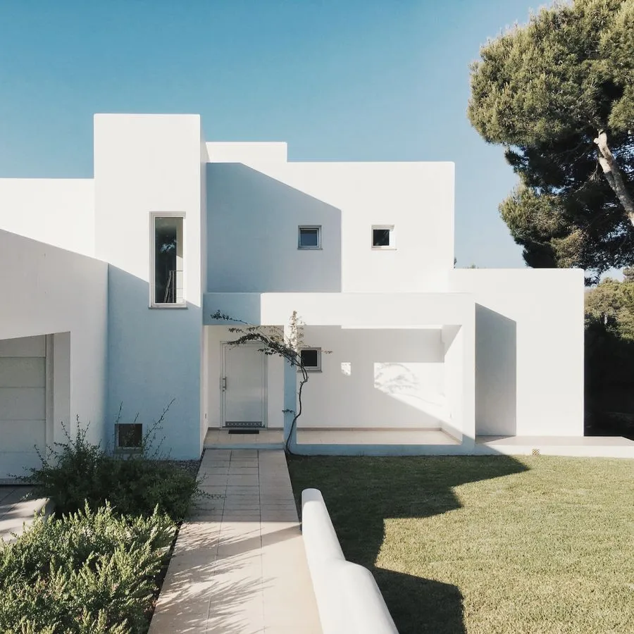 A white rendered modern house with a walled courtyard, lawn and a large pine tree