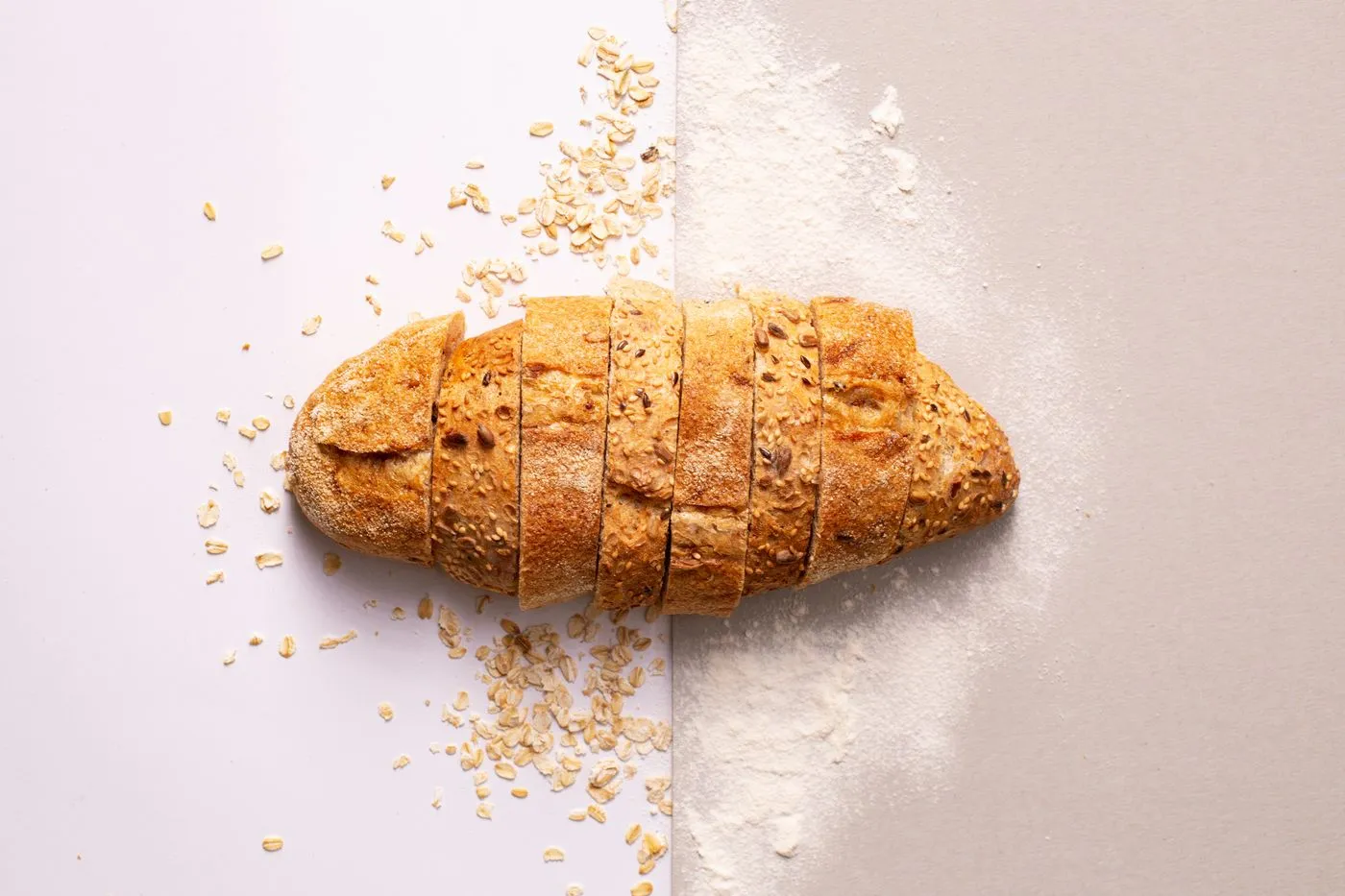 Sliced seeded bread surrounded by oats on a flour-dusted surface